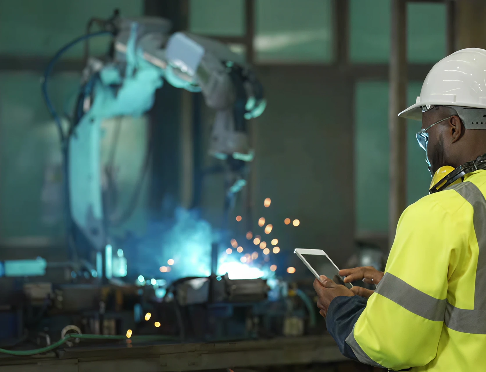 A worker in protective gear monitors a robotic welding cell from a tablet on a production floor.